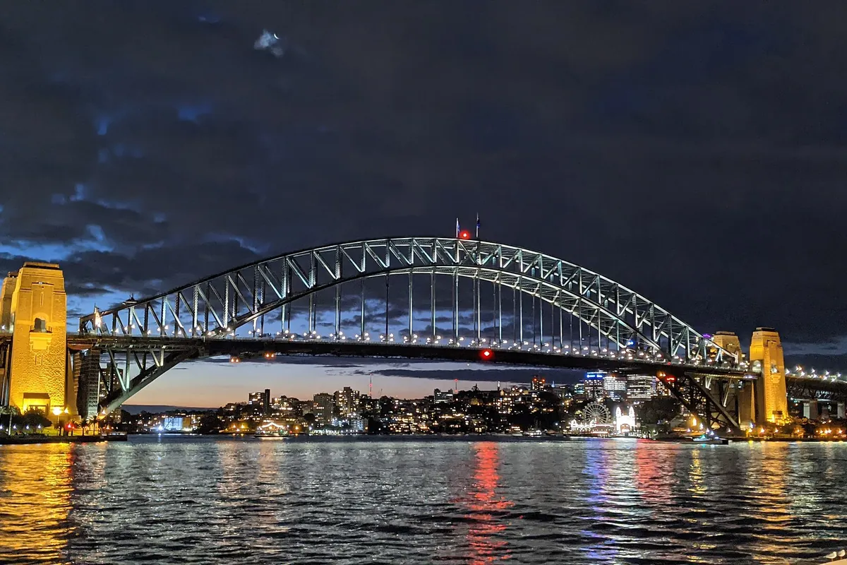 The Sydney Harbour Bridge at night, its arch and floodlit sandstone pylons reflected in the harbour, with the lights of the north shore behind.