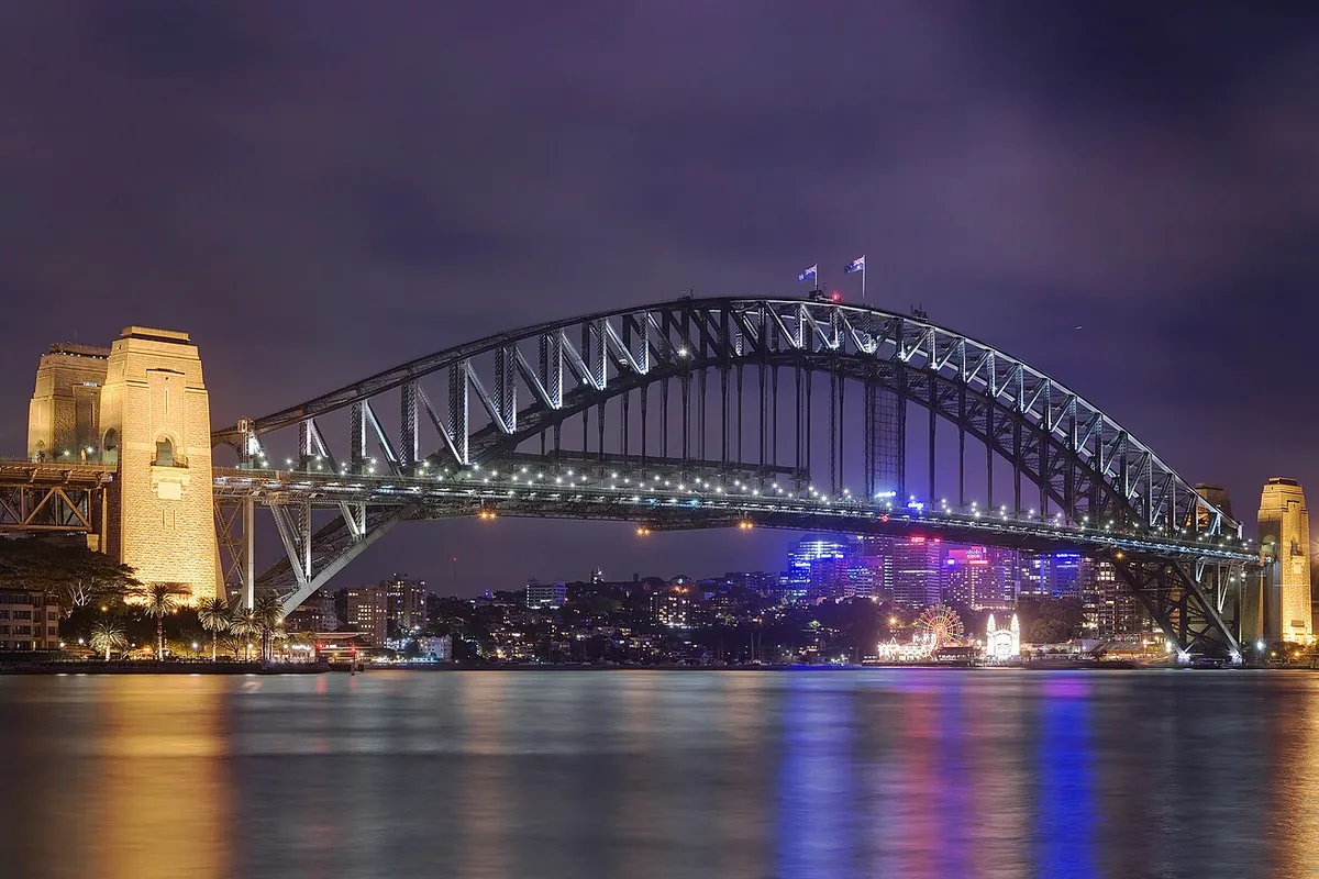 The Sydney Harbour Bridge at night seen from Circular Quay, its arch picked out in lights against a purple sky with a sandstone pylon on the left.