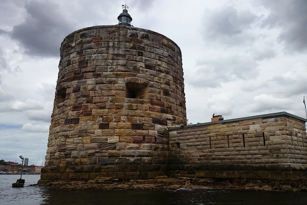 The round sandstone Martello tower of Fort Denison seen from the water, with a lantern on top and a low stone wall running to the right.
