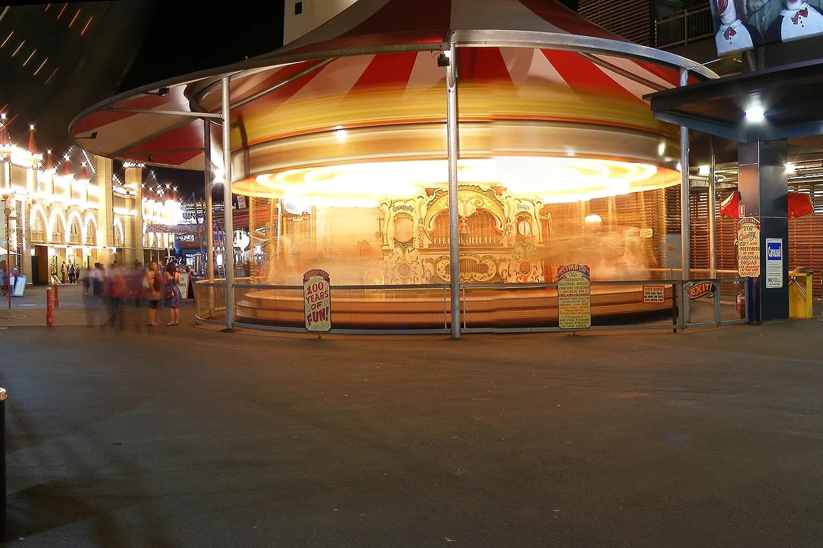 A carousel with a red-and-white striped canopy glowing at night at Luna Park Sydney, with the strung lights of a building to the left.