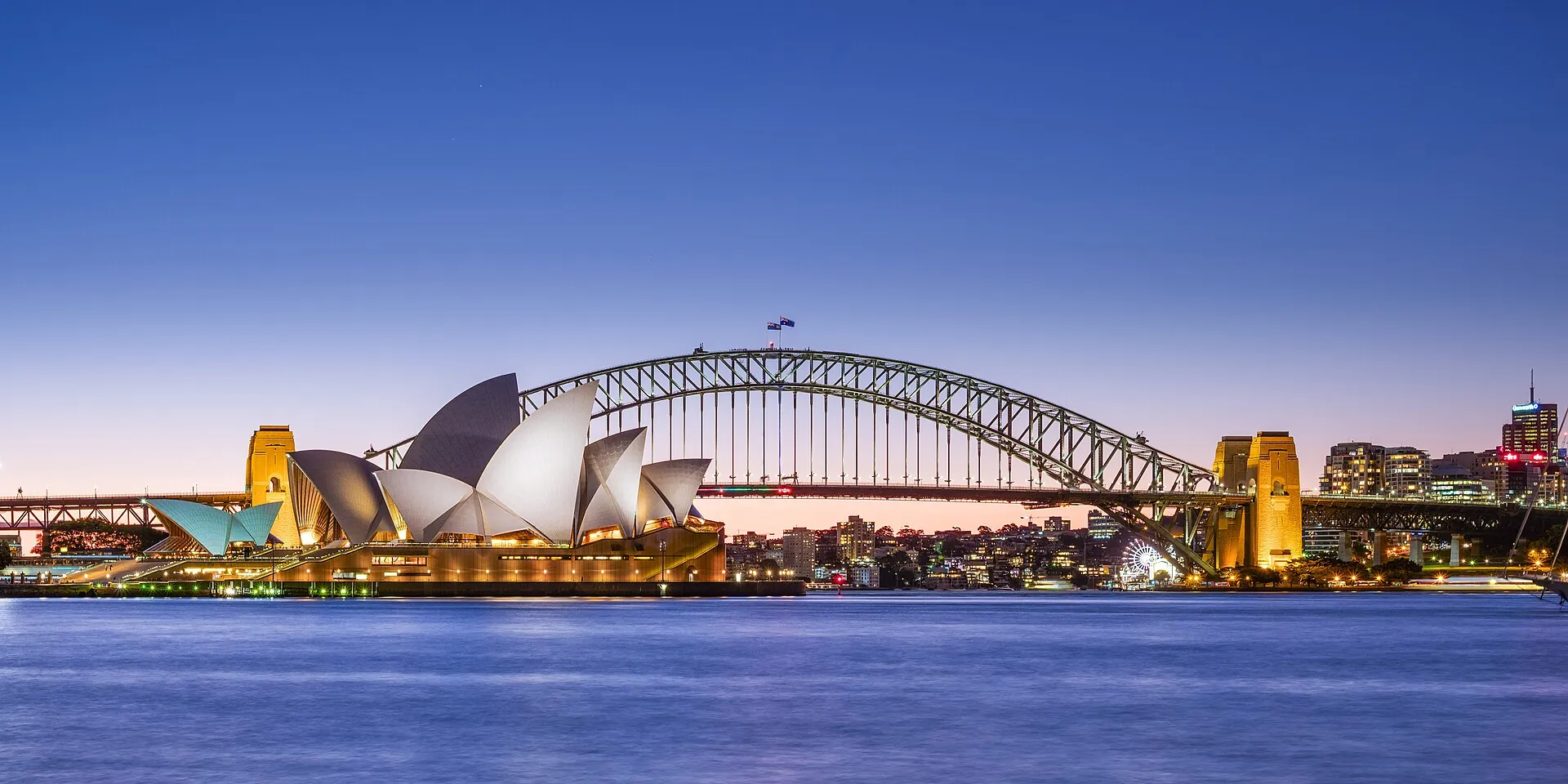 The Sydney Opera House and the Sydney Harbour Bridge at dusk, with the lit sails of the Opera House and the steel arch of the bridge across calm blue water.