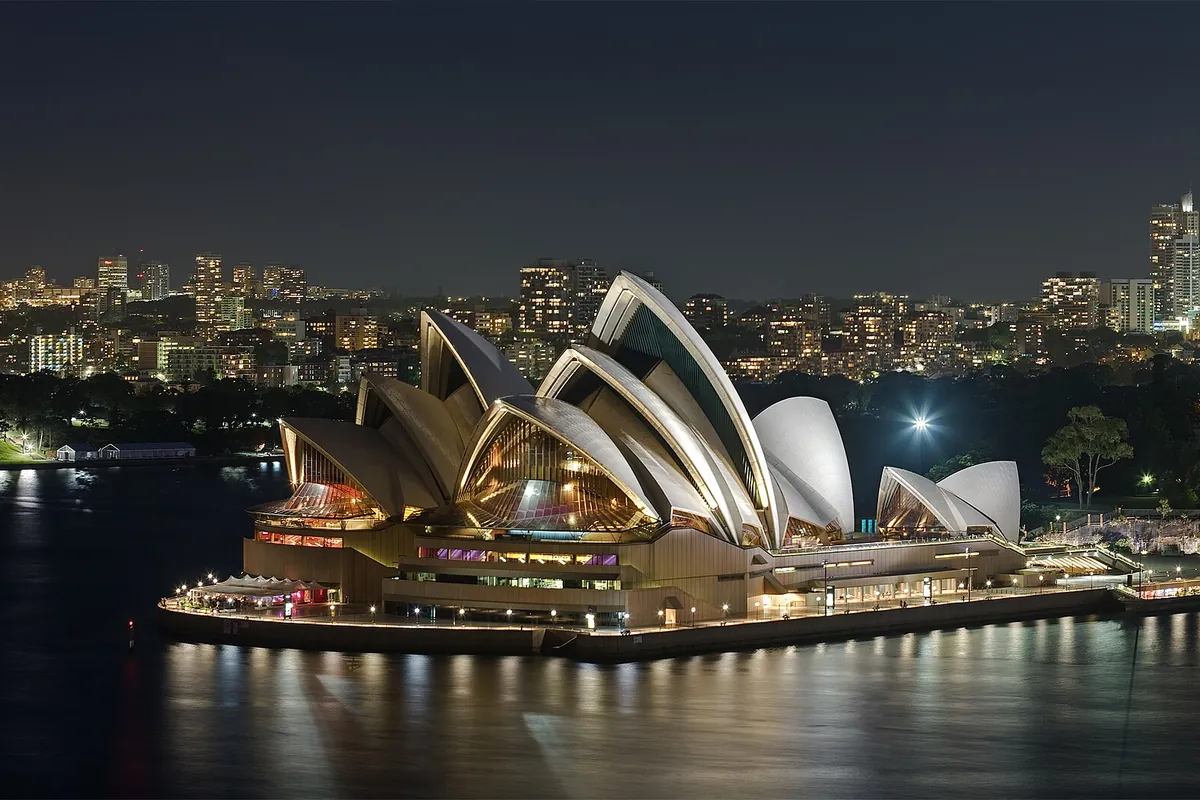The Sydney Opera House lit at night, its white sails and glass ends glowing above the dark harbour, with the lights of the city behind.
