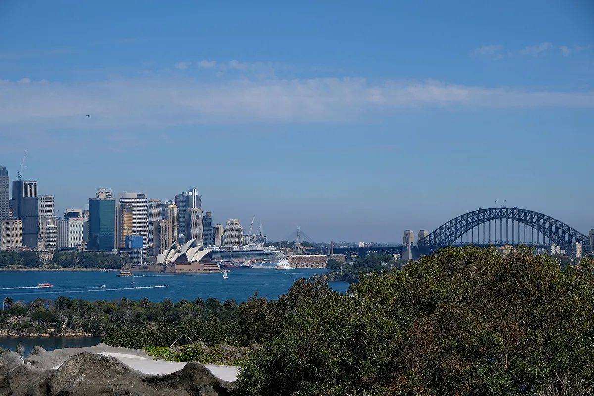 A view across Sydney Harbour from the Taronga Zoo shore to the city towers, the Opera House and the Harbour Bridge, with green bushland in the foreground.